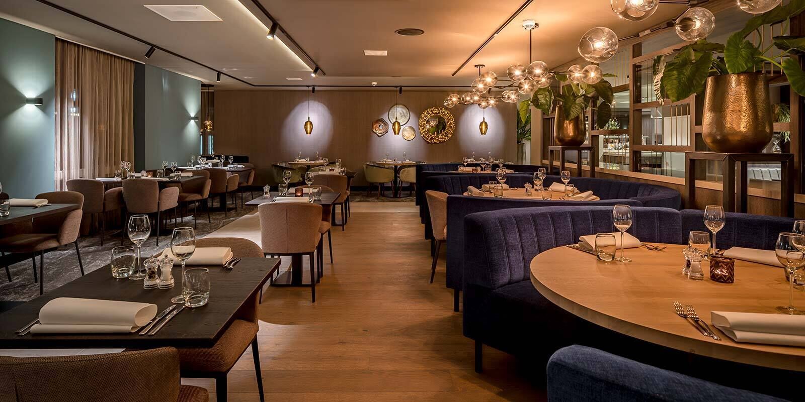 Un restaurant avec des tables rondes, des banquettes en tissu bleu foncé et une ambiance chaleureuse et accueillante, agrémentée de luminaires suspendus et de plantes.