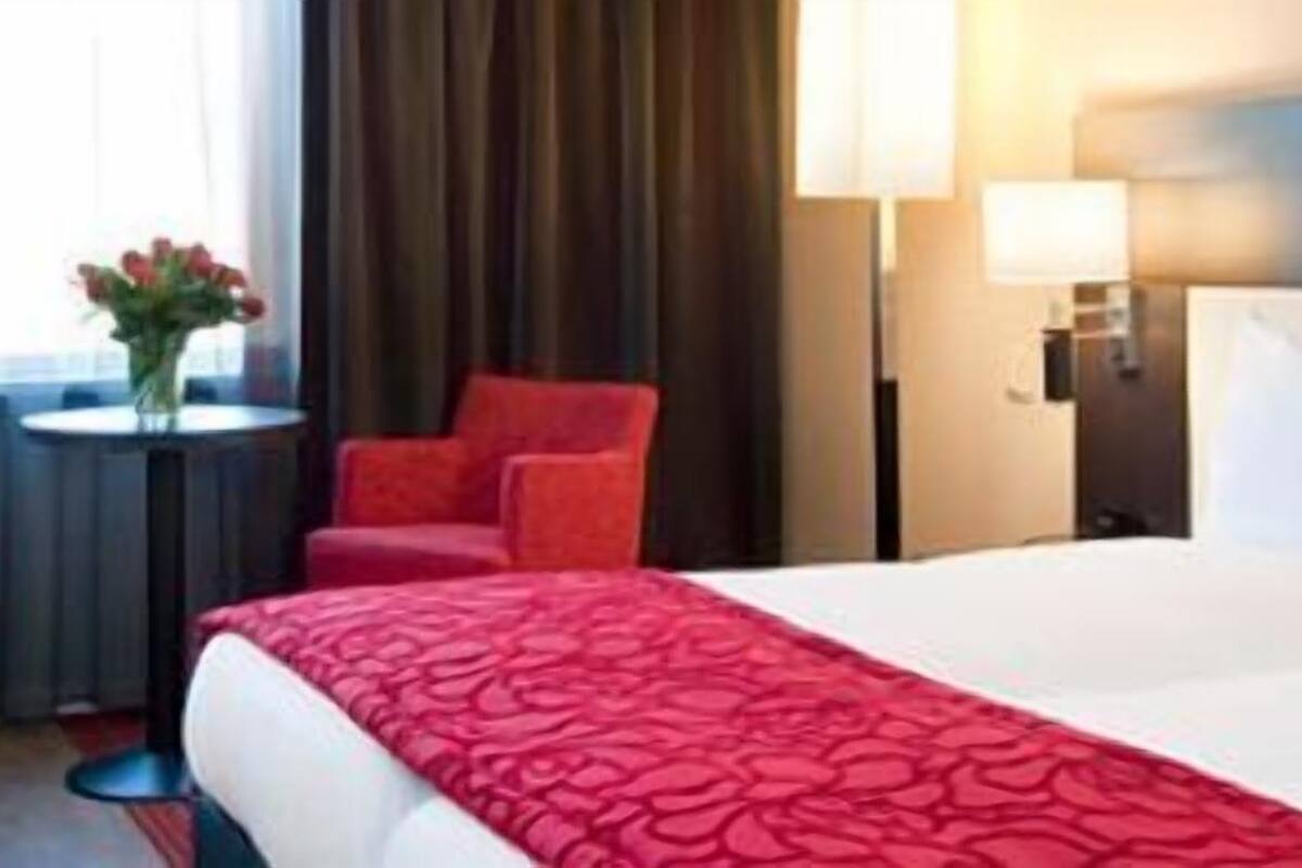 A hotel room with a large bed, a desk with a laptop, two red chairs, and a small table with flowers.
