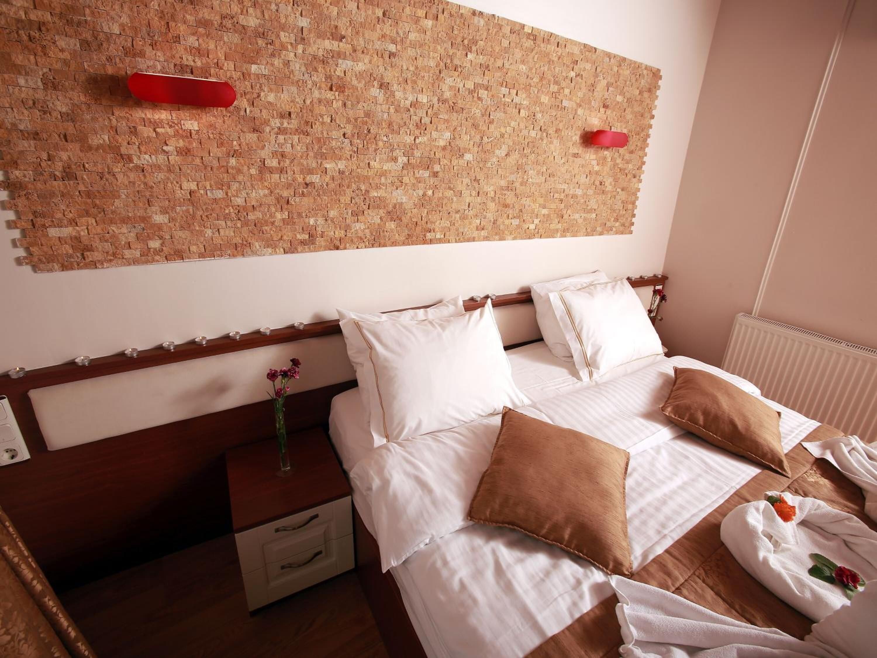 A hotel room with a brick-patterned wall, a wooden headboard, a bedside table with a vase of flowers, and a neatly made bed with white and brown pillows.
