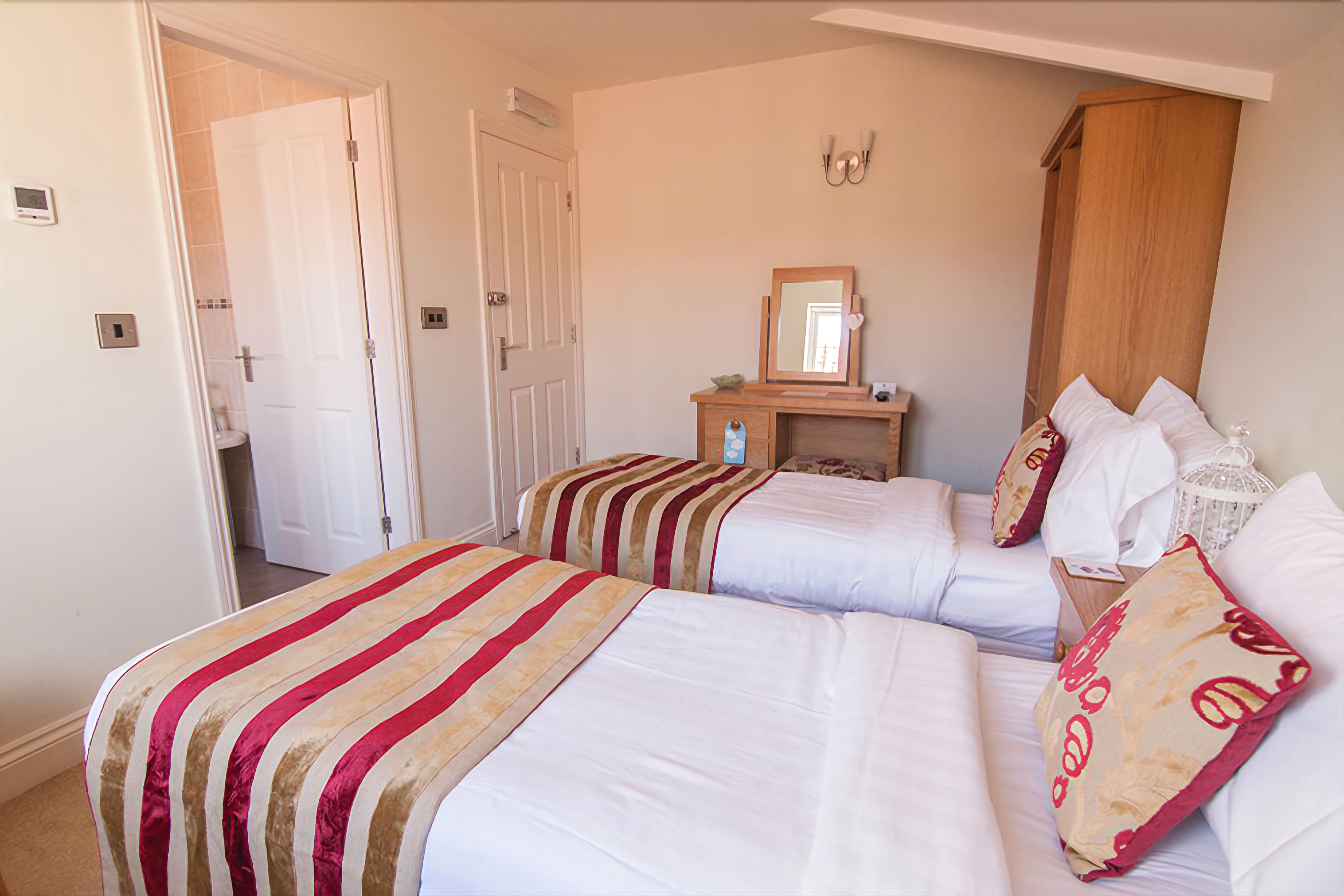 A hotel room with two beds, a wooden wardrobe, a bedside table with a mirror, and a nightstand with a lamp.