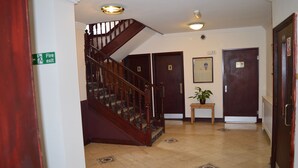 A hallway with a wooden staircase, a fire exit sign, and a potted plant. - The Globe Hotel (Colchester)