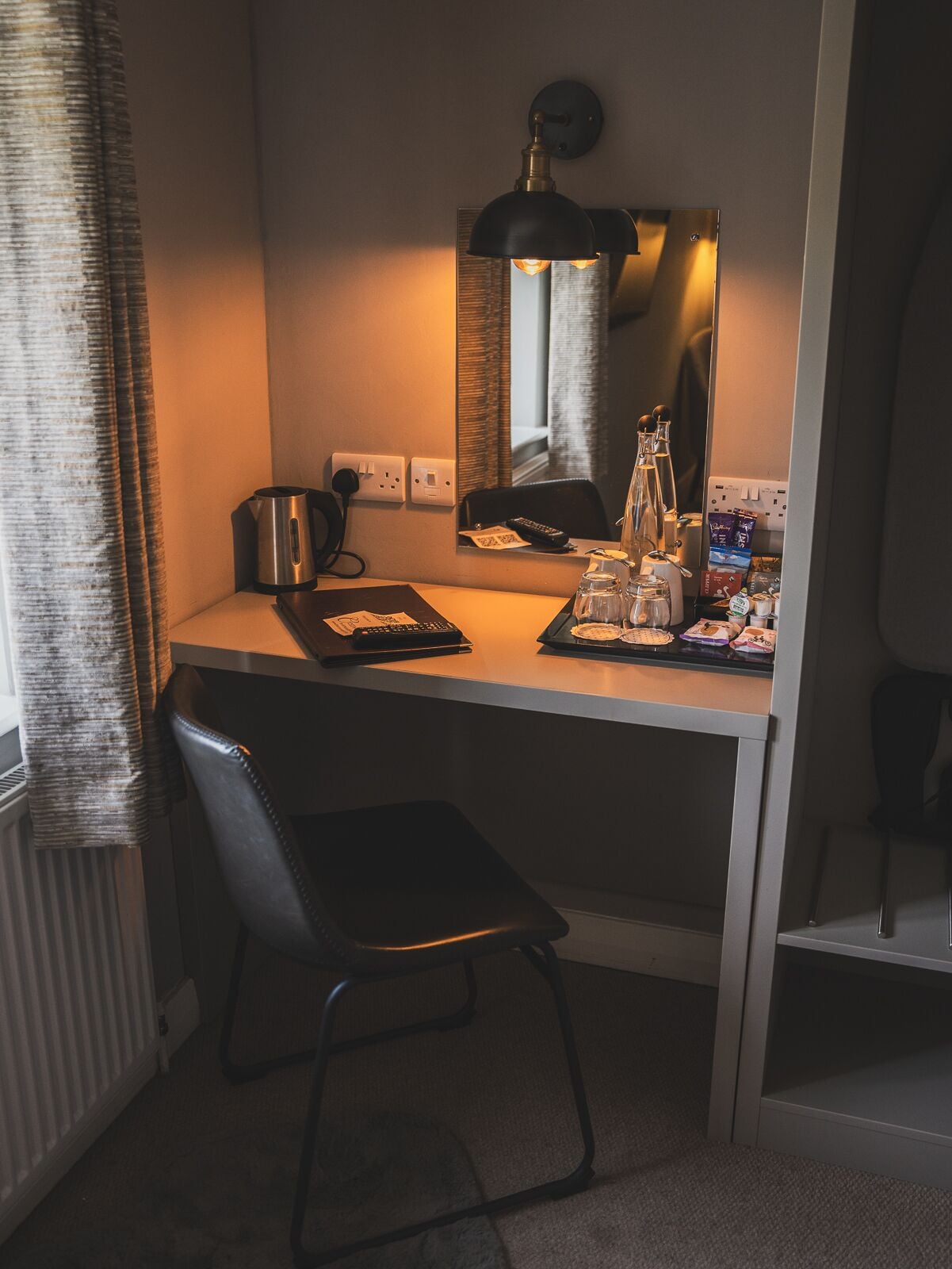 A small, modern hotel room with a compact desk, a chair, a kettle, a mirror, and a window with curtains.