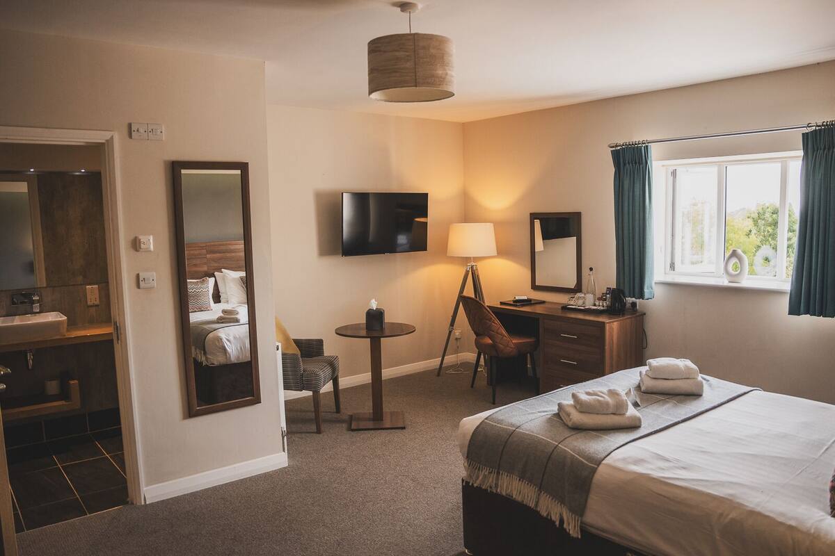 Habitación de hotel con cama, escritorio, silla, televisor, lámpara y ventana con cortinas.