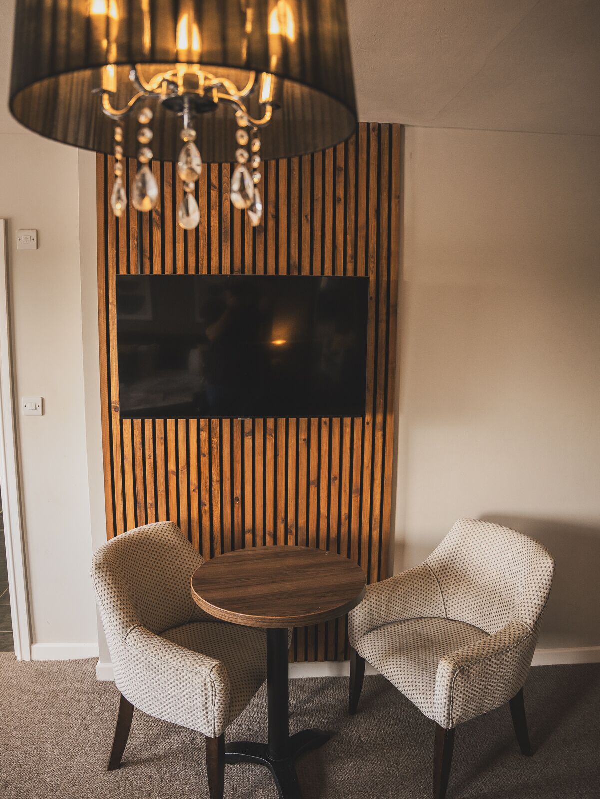 A room with a round wooden table, two patterned chairs, a mounted flat-screen TV, and a wooden slat wall.
