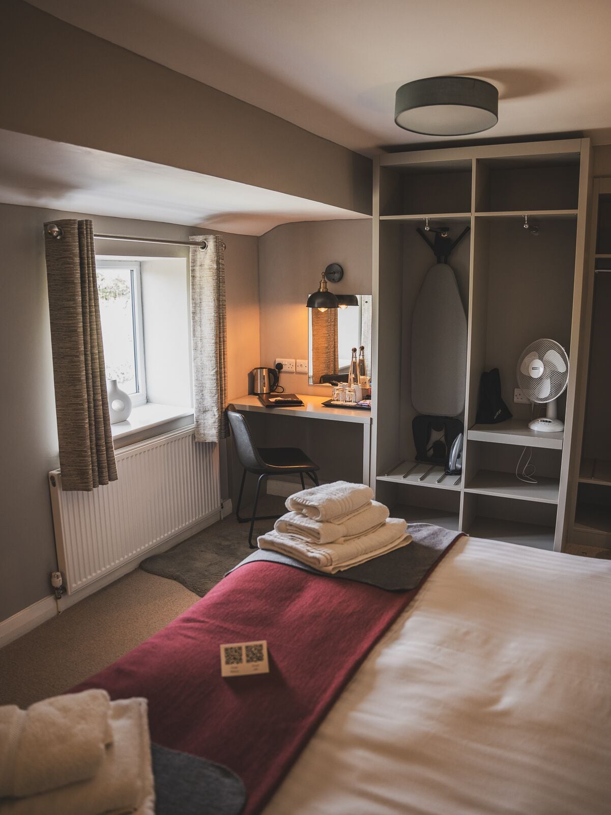 Habitación de hotel con cama, escritorio con silla, armario, radiador y ventana con cortinas.