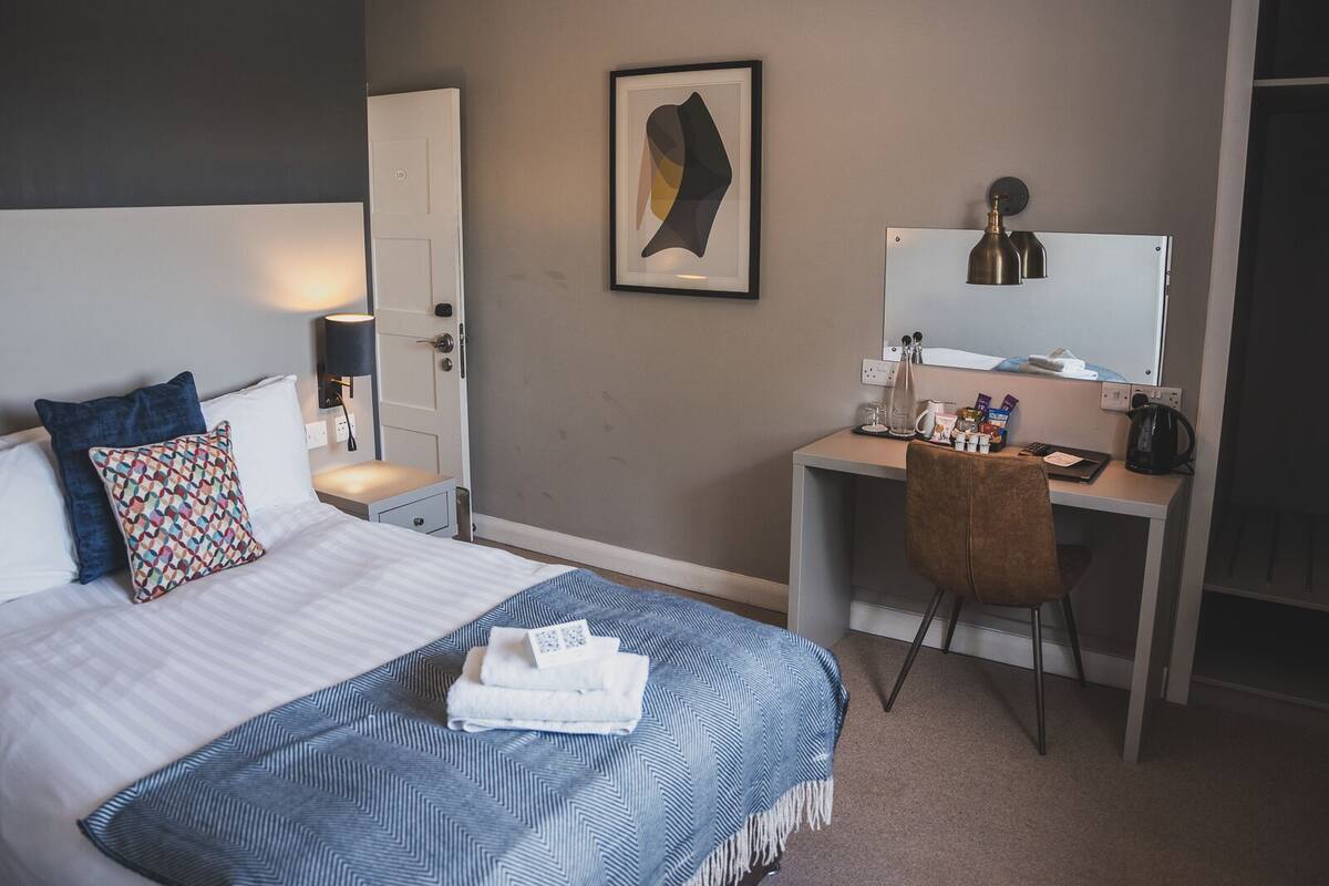 Habitación de hotel con cama, escritorio, silla, espejo y una obra de arte colgada en la pared.