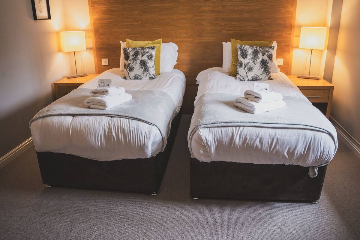 Dos camas individuales con cabeceros de madera, ropa de cama y toallas blancas. Cada cama tiene una mesita de noche con una lámpara y un cuadro en la pared.