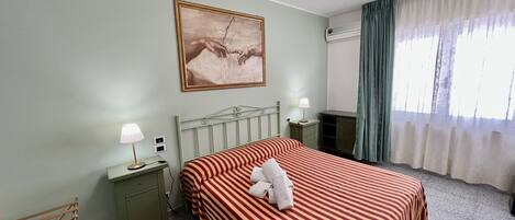 A hotel room with a bed, a nightstand, a lamp, a painting on the wall, and a window with curtains.