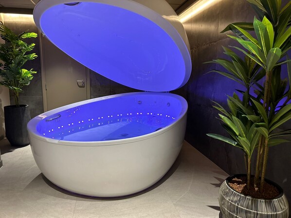 Una tina de hidromasaje grande, blanca y ovalada, con interior azul y luces LED, rodeada de macetas con plantas.