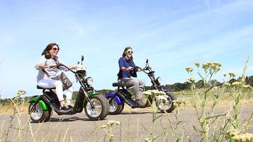 Two people riding electric scooters on a paved road with grass and wildflowers on the sides.