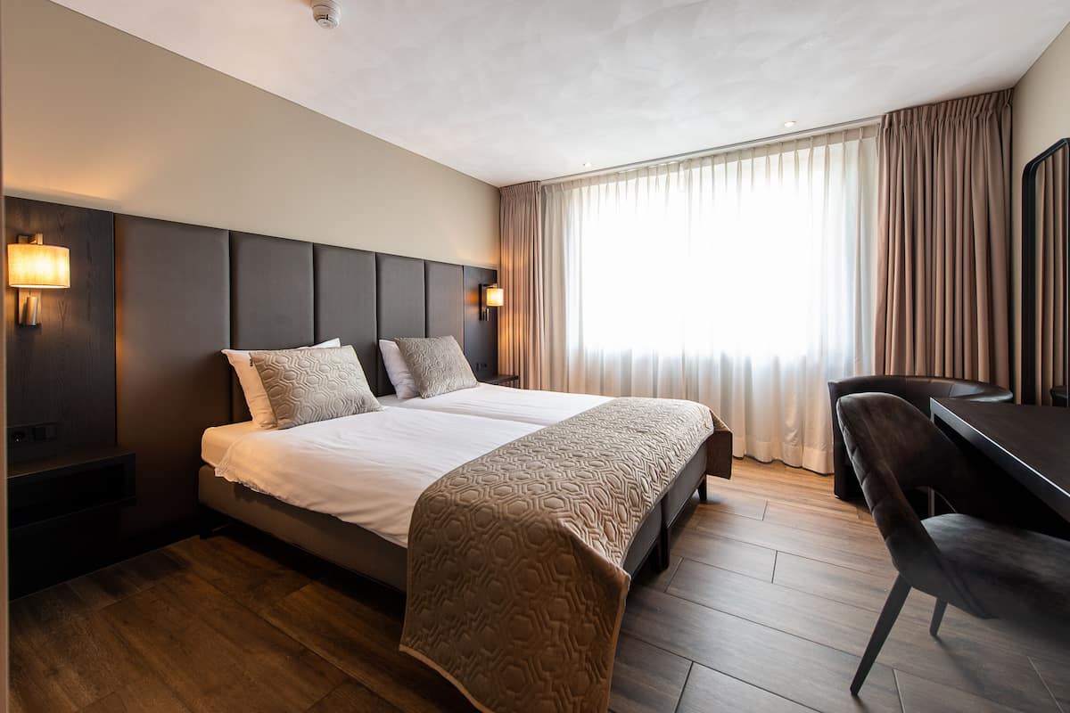 Une chambre d’hôtel avec un grand lit, des lampes de chevet, un bureau et une vue sur la ville à travers de fines rideaux.