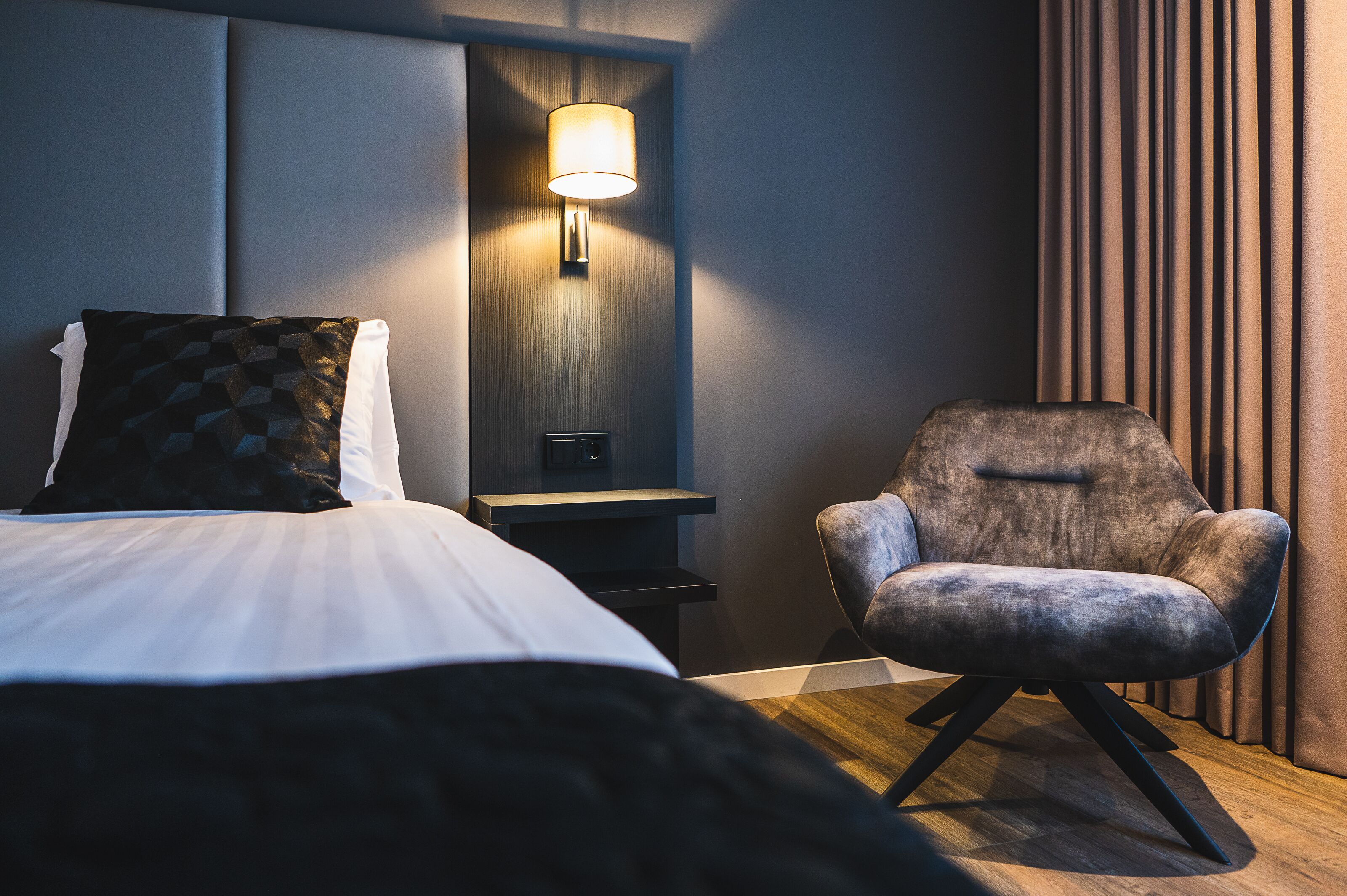 Ein modernes Hotelzimmer mit Bett, Sessel, Nachttisch und Wandlampe.