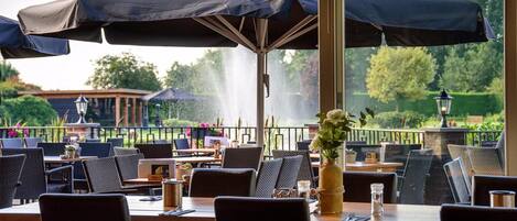 An outdoor dining area with tables and chairs, umbrellas, and a view of a garden and trees.