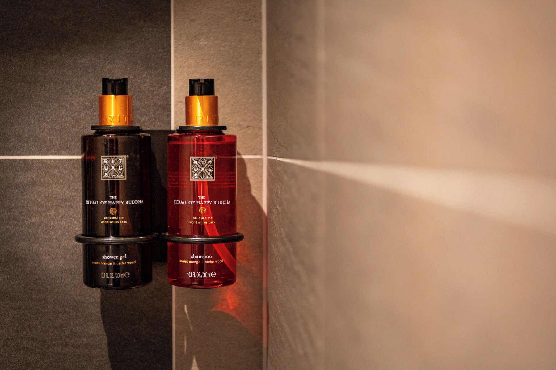 Zwei Flaschen Ritual of Happy Buddha Produkte, eine als Duschgel, die andere als Shampoo gekennzeichnet, beide mit einem süßen Duft von Orange und Zedernholz.
