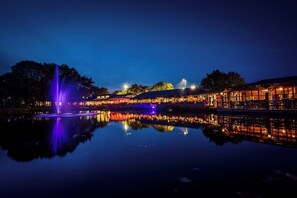 Hotel frente a un lago con una fuente, iluminado con luces moradas, y su reflejo en el agua.