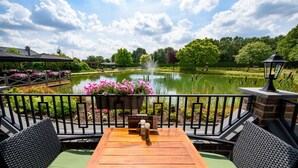 A wooden table with wicker chairs, a sign for lunch and drinks, and a view of a pond with flowers and trees. - Maashof (Venlo)