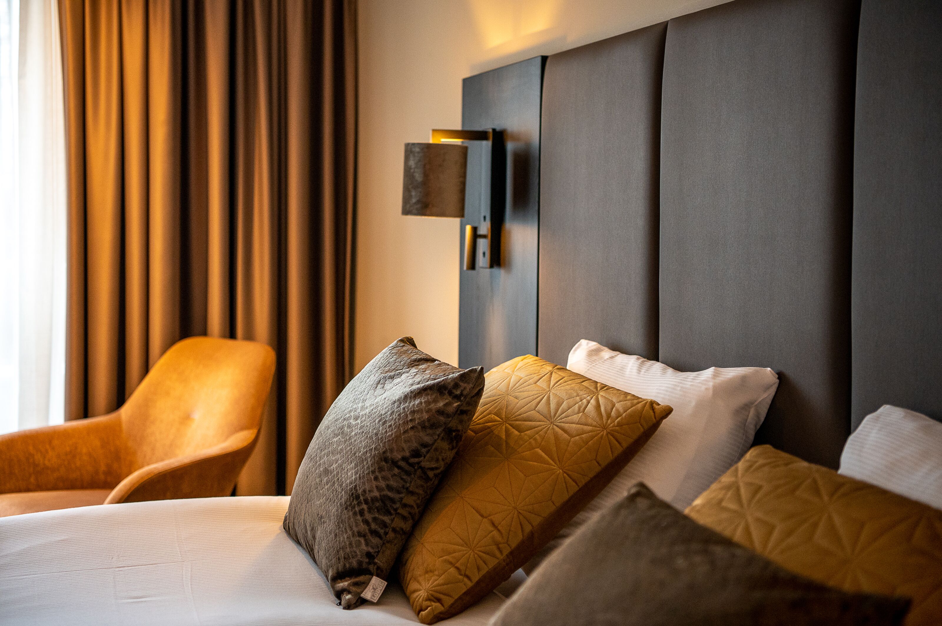 A modern hotel room with a large bed, a wooden armchair, and a wall-mounted light fixture.
