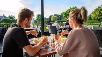 Een stel eet aan een buitentafel, met uitzicht op een tuin en een gebouw op de achtergrond.