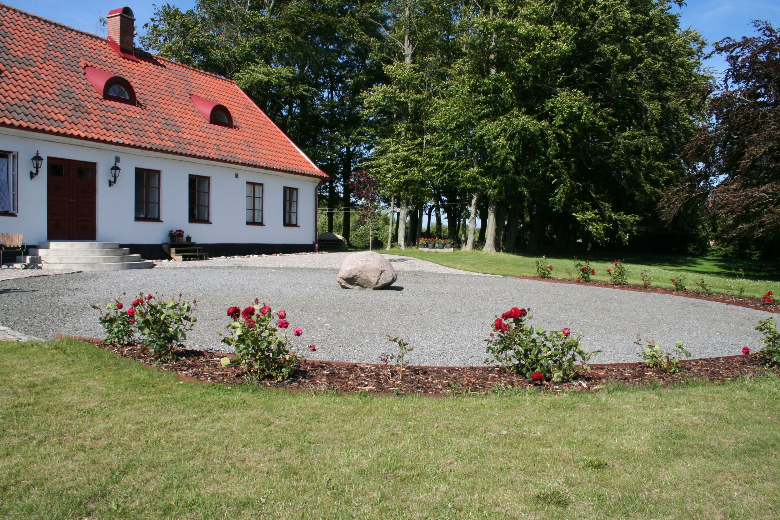 En vit byggnad med ett rött tak, omgiven av en grusuppfart och en blomsterrabatt med röda rosor.