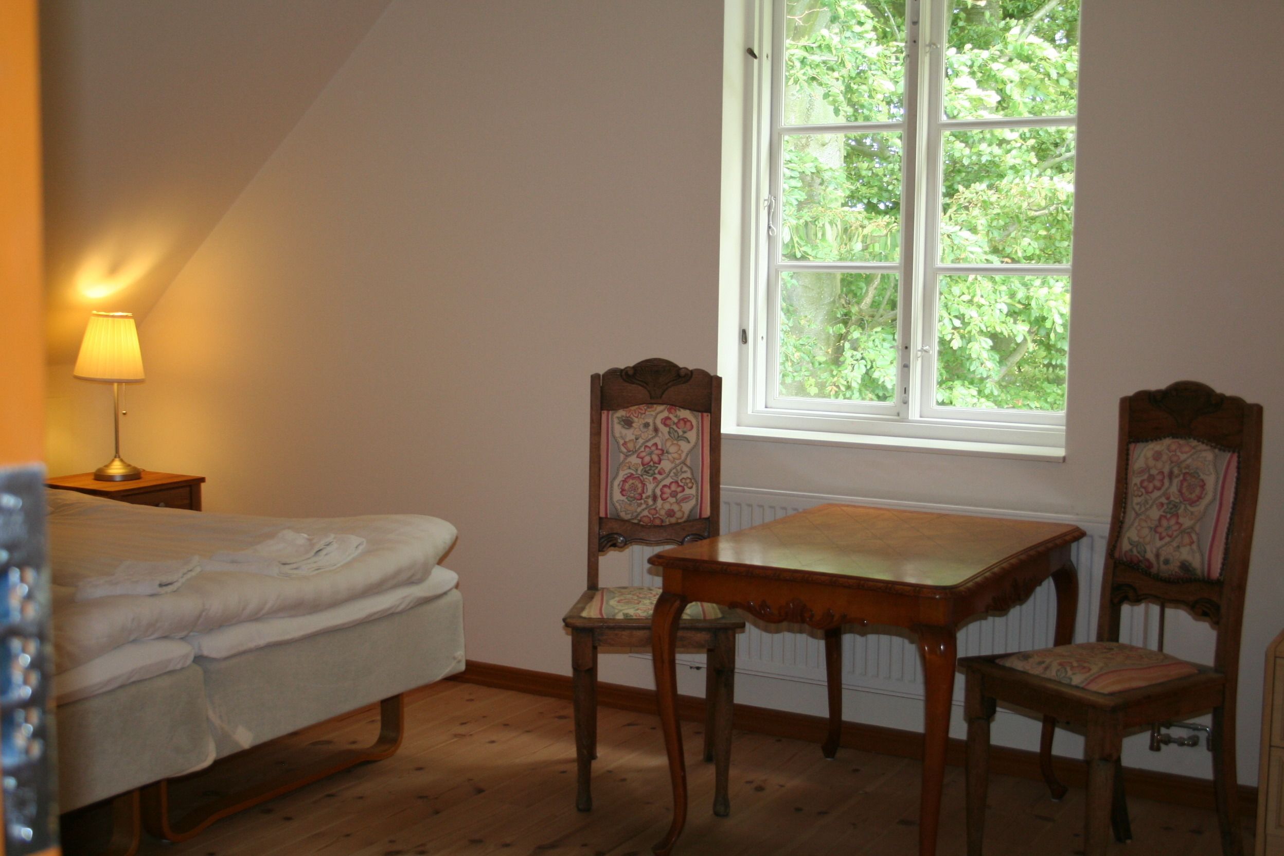 A bedroom with a bed, a table, two chairs, a lamp, and a window with a view of trees.