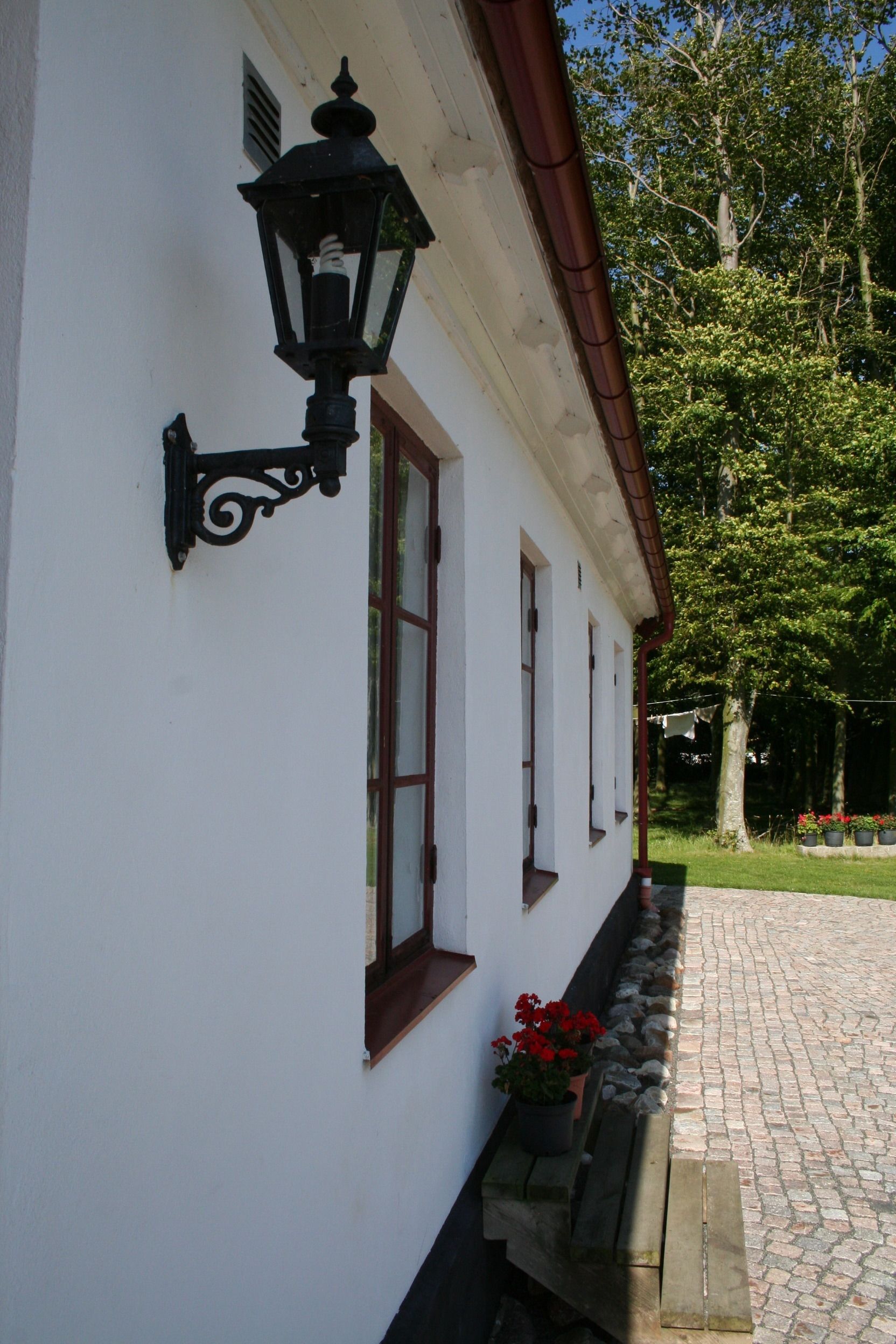 Un bâtiment blanc avec une lampe noire fixée au mur, une fenêtre avec un cadre en bois et un banc en pierre agrémenté de fleurs rouges.