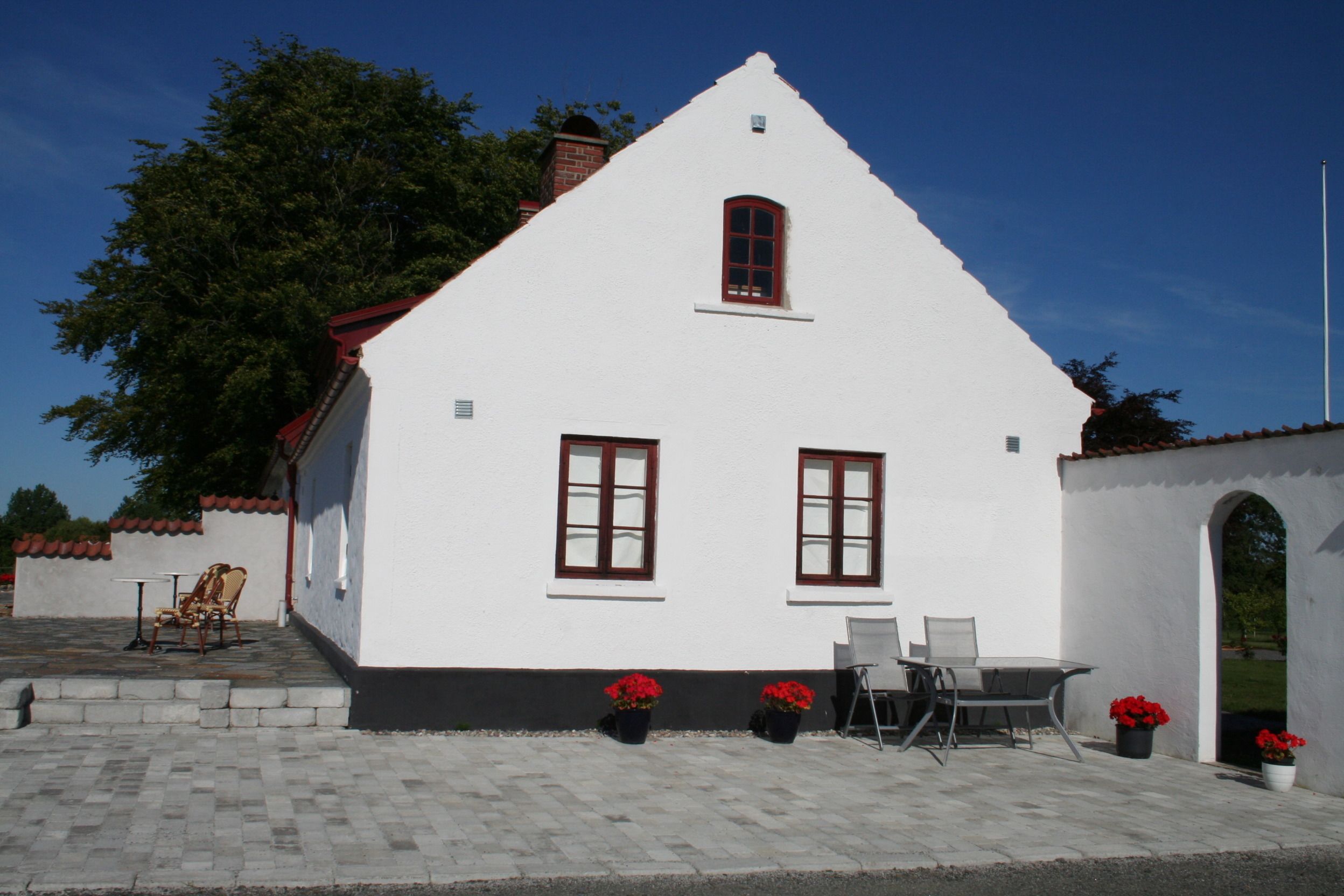 A white building with a red roof, two windows, and a door. There are potted plants and outdoor furniture.