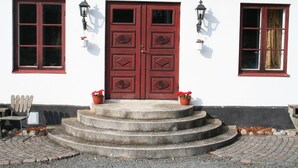 Un bâtiment blanc avec une porte rouge à deux battants, deux fenêtres aux encadrements rouges et un escalier en pierre menant à la porte.