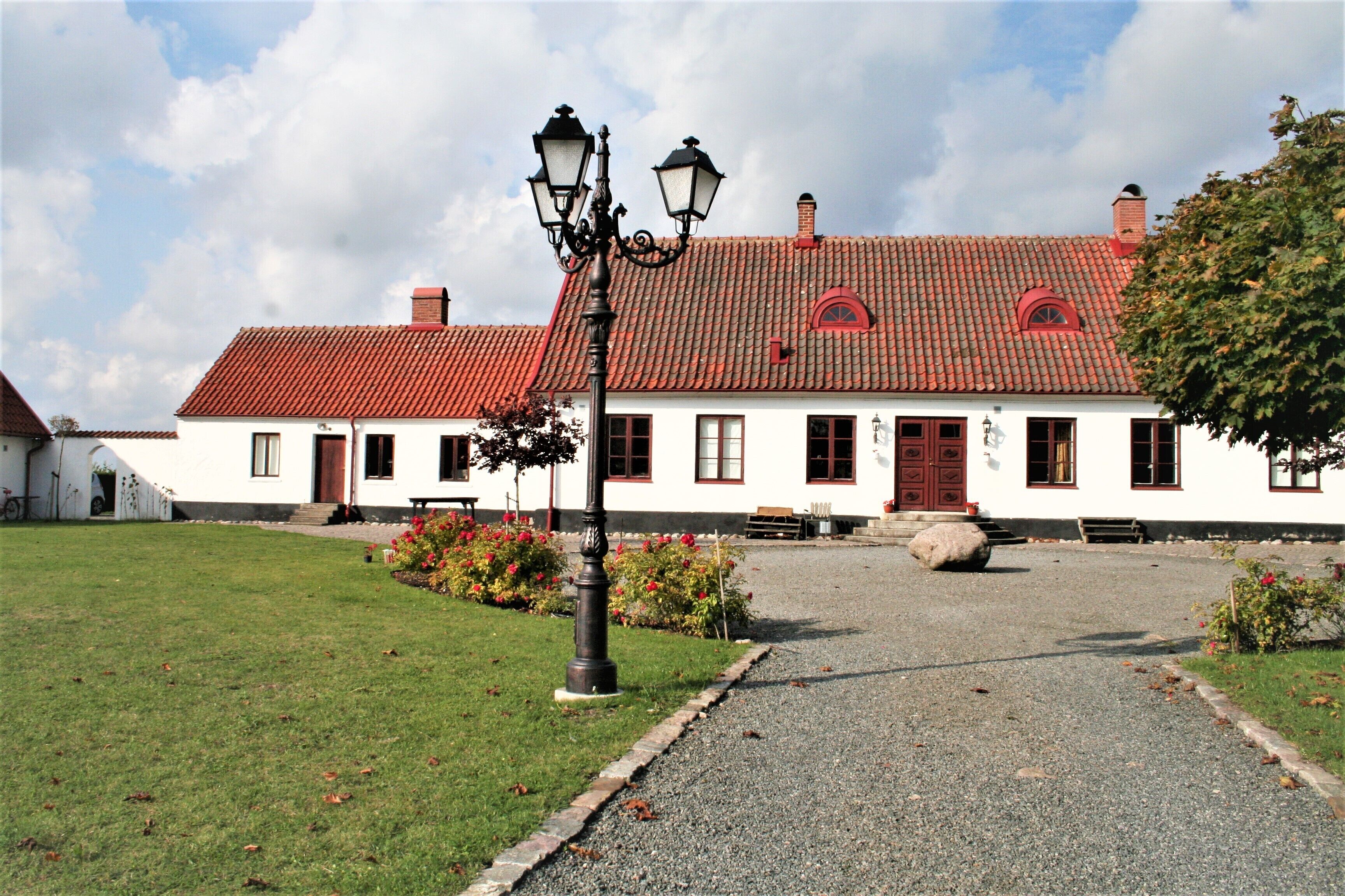 En vit byggnad med ett rött tegeltak, en grusgång som leder till entrén och en gatlykta i förgrunden.