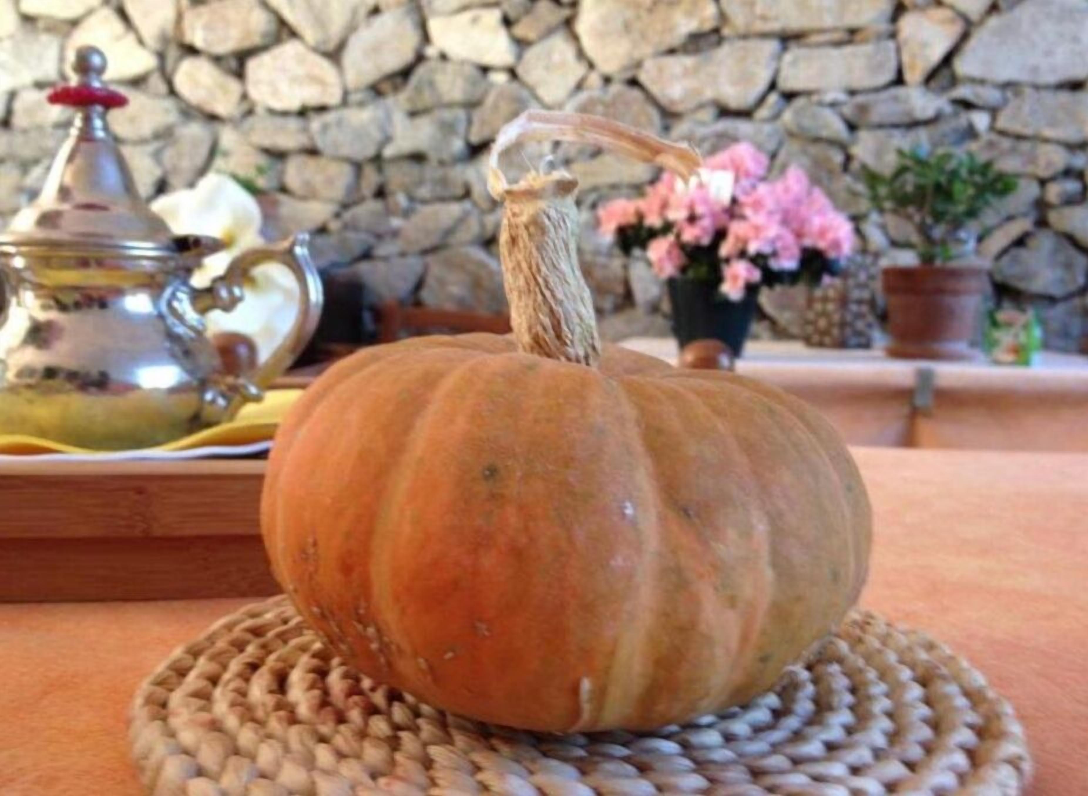 A pumpkin on a woven placemat with a stone wall and a metal teapot in the background.