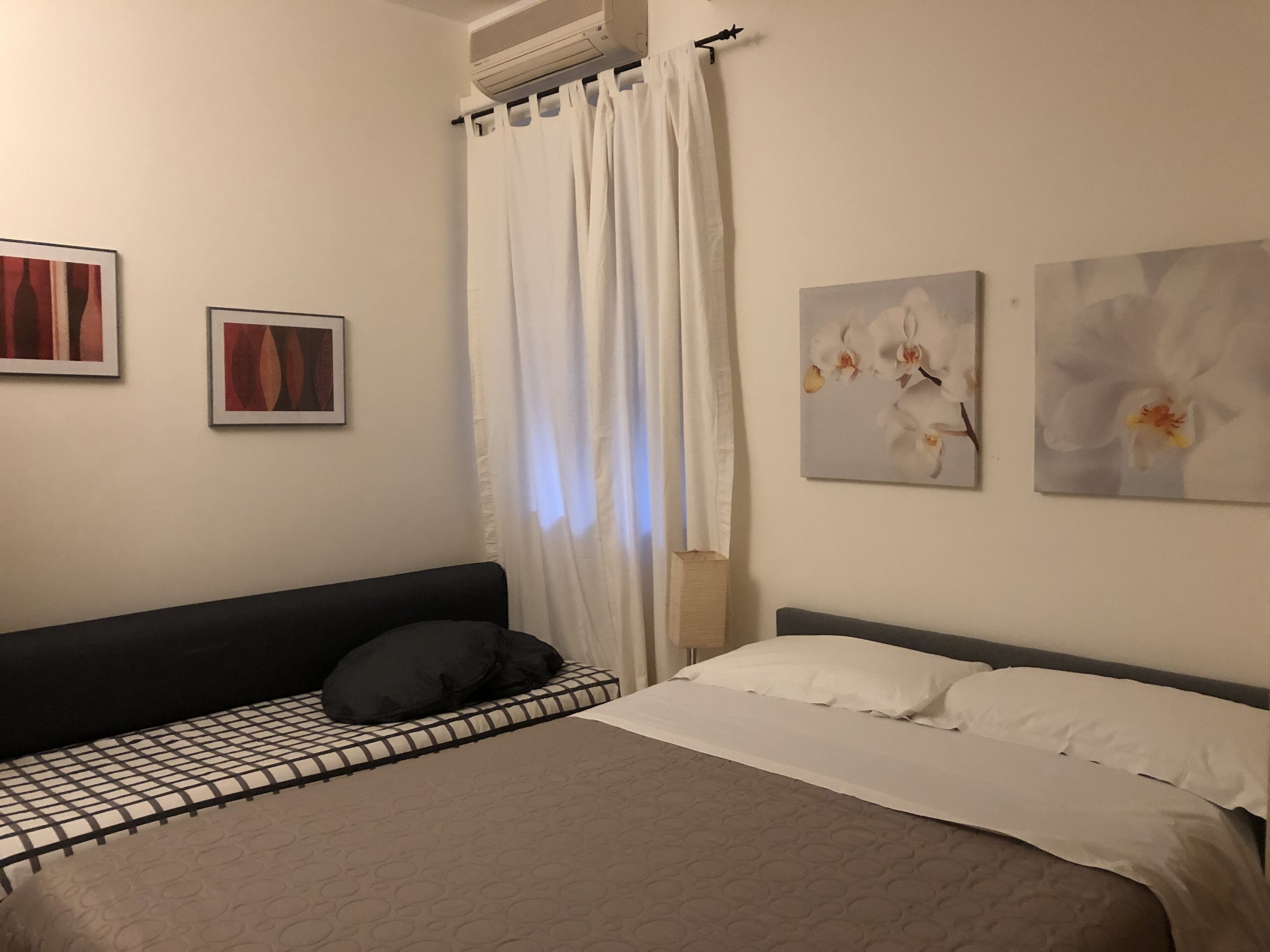 Un dormitorio con una cama, una mesita de noche, un aire acondicionado de pared y dos cuadros enmarcados.