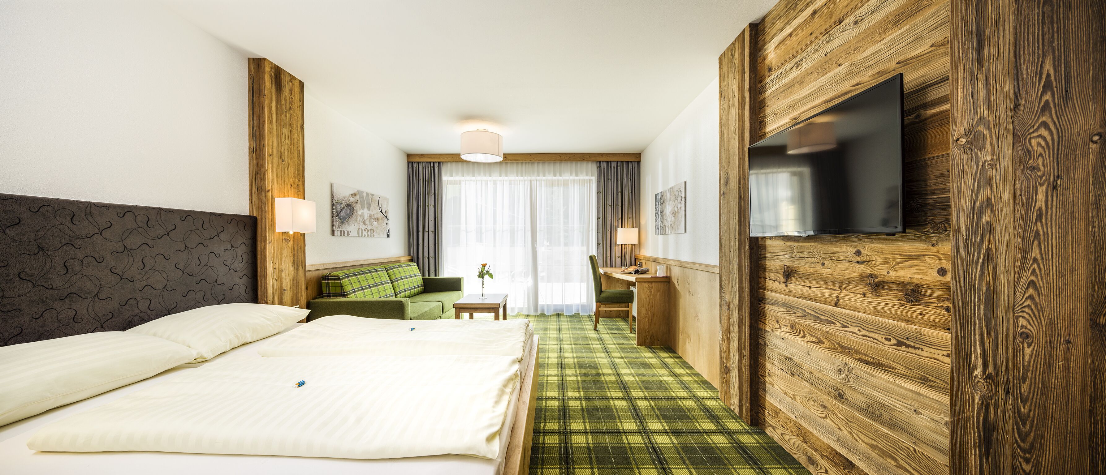 Quarto de hotel com cama, sofá verde, escrivaninha, televisão e parede de destaque em madeira.