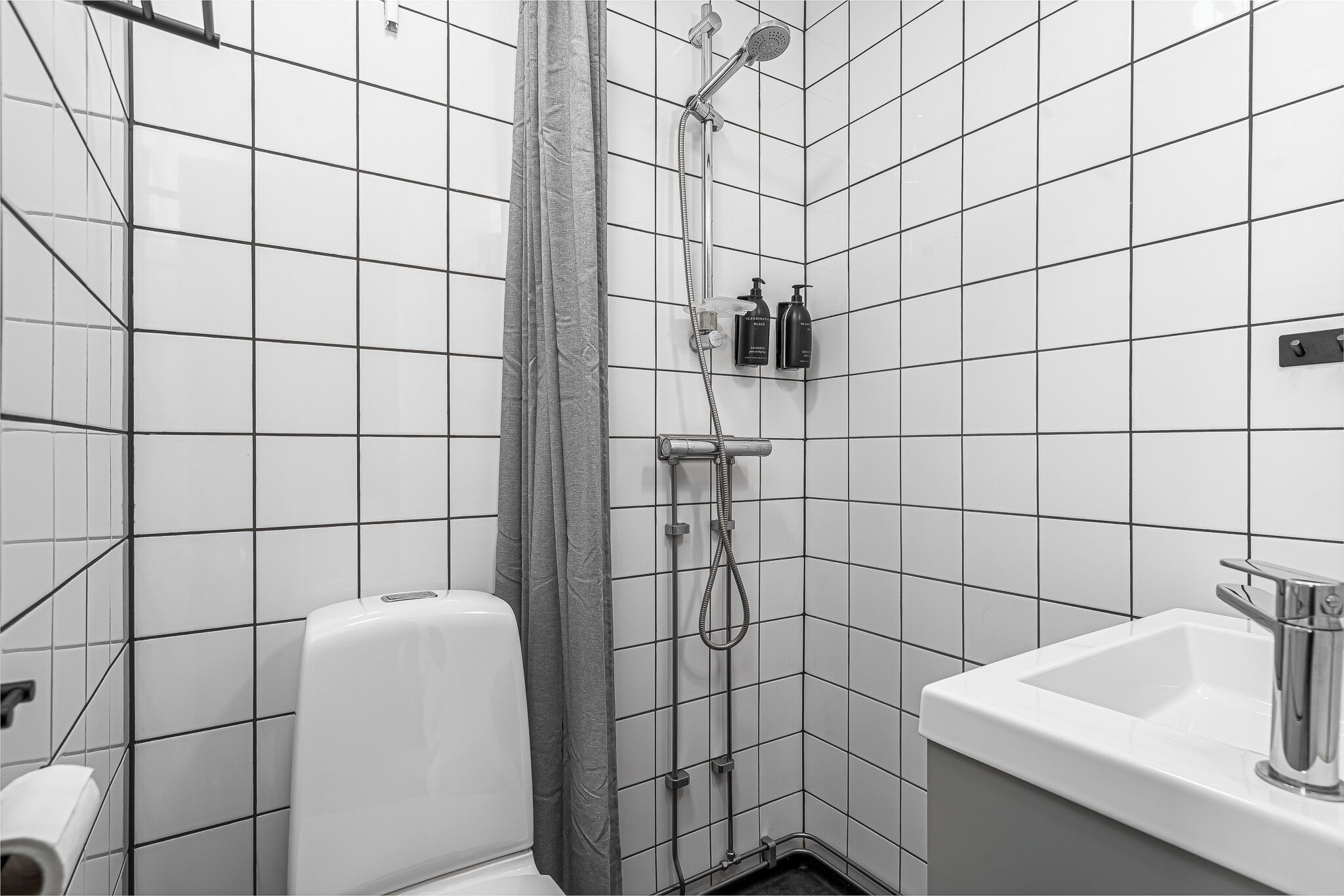 Un baño con paredes de azulejos blancos, una ducha con cortina, un inodoro y un lavamanos con grifo.