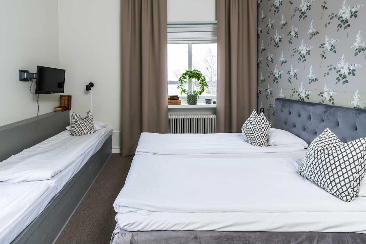 A hotel room with two single beds, a large bed, a TV on the wall, a window with curtains, and a radiator.
