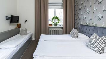 Habitación de hotel con dos camas individuales, una cama grande, un televisor en la pared, una ventana con cortinas y un radiador.