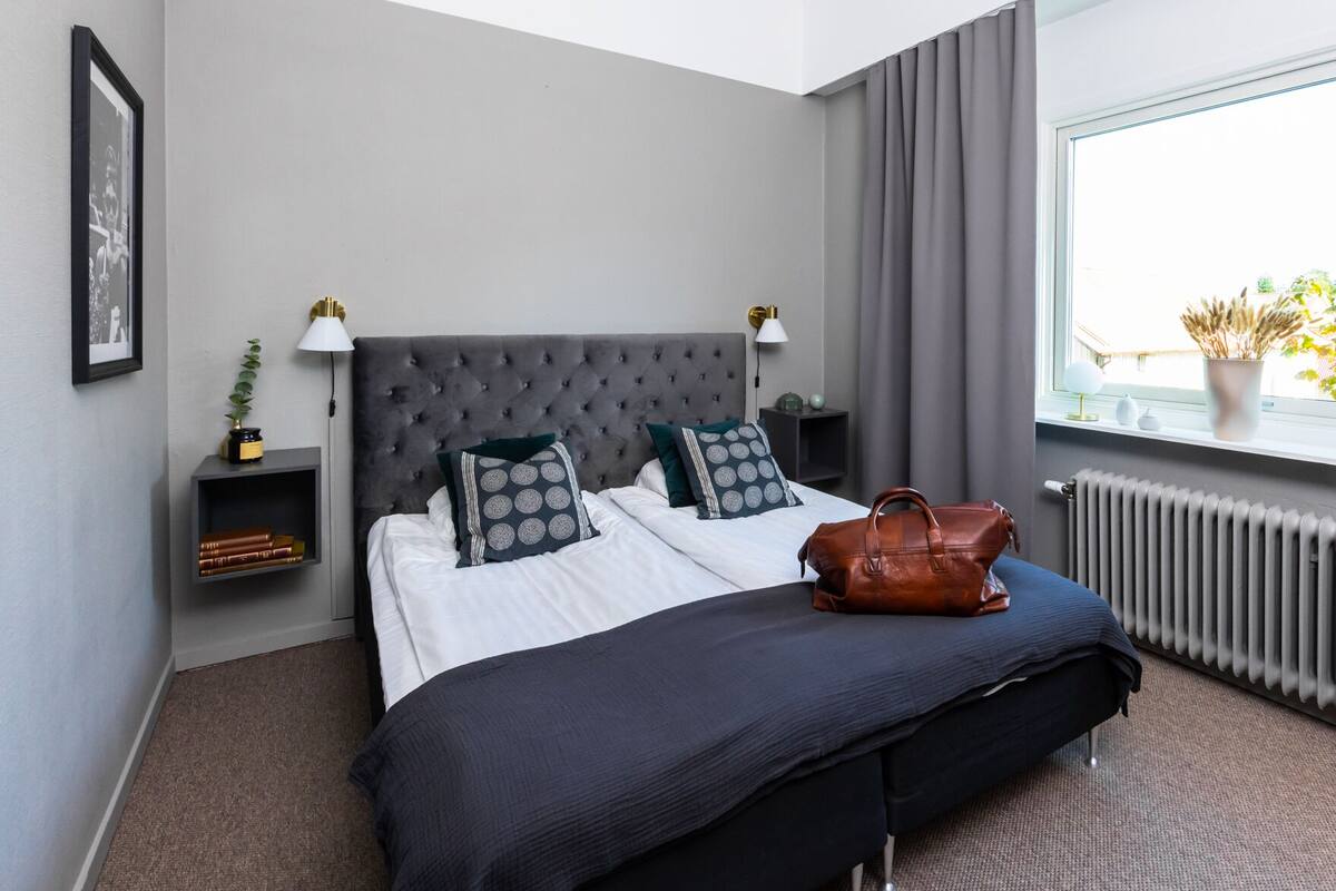 A neatly made bed with a grey headboard, two bedside tables with lamps, a window with a view of greenery, and a radiator.
