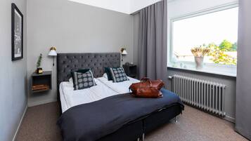 A neatly made bed with a grey headboard, two bedside tables with lamps, a window with a view of greenery, and a radiator.