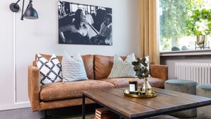 A living room with a brown leather sofa, a wooden coffee table with books, a black and white photograph on the wall, and a window with a plant. - Stora Hotellet Markaryd (Markaryd)