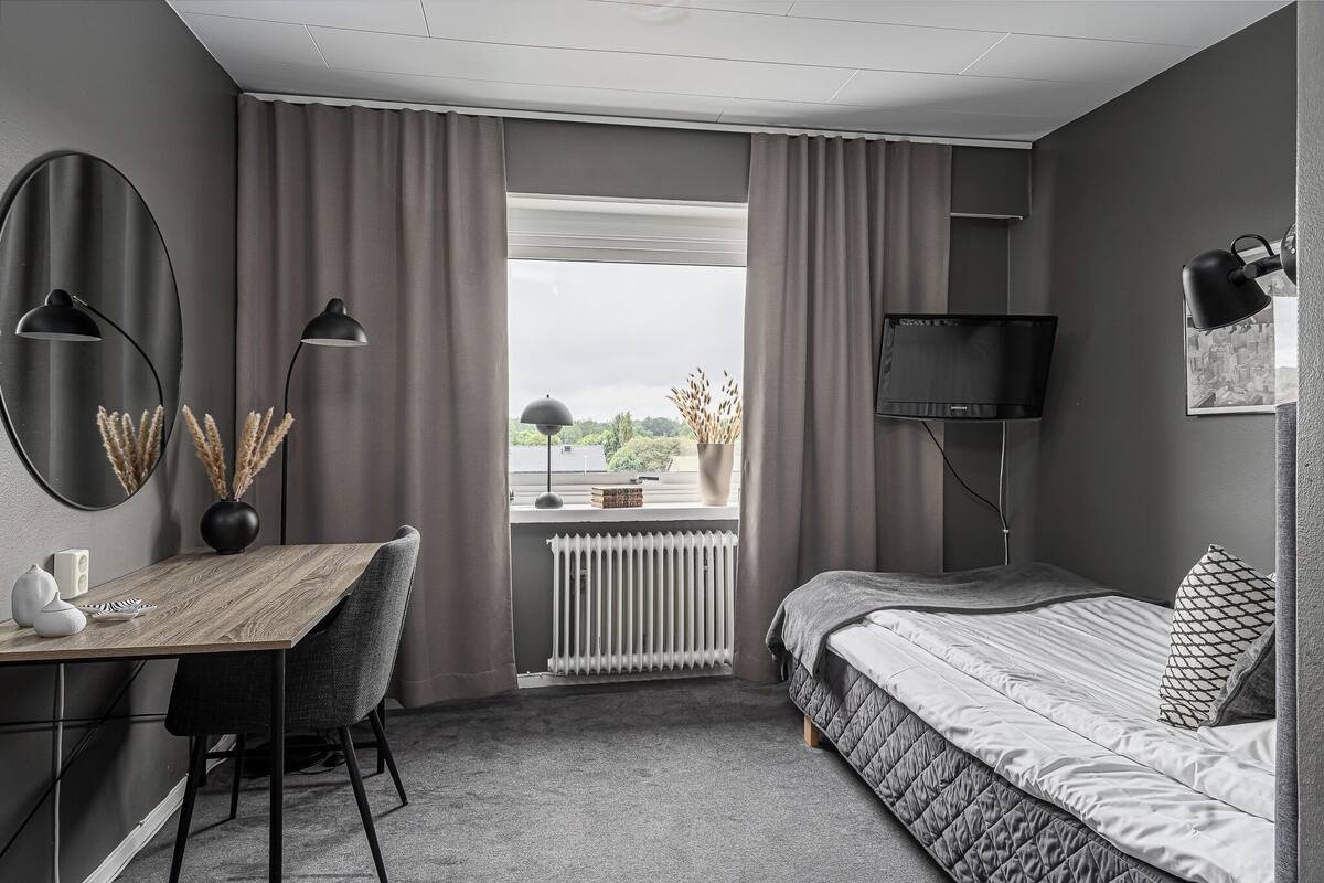 A bedroom with a bed, a desk, a chair, a radiator, a window with curtains, and a TV.