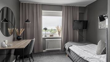 Een slaapkamer met een bed, een bureau, een stoel, een radiator, een raam met gordijnen en een televisie.