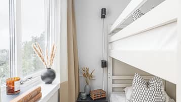 A bunk bed room with a window, a bedside table with a candle, a vase with dried plants, and a patterned pillow.