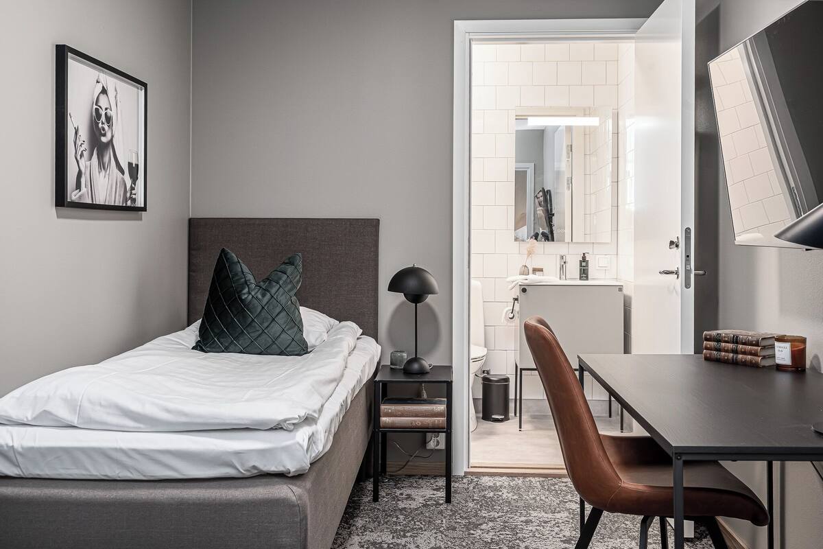 A bedroom with a bed, bedside table, lamp, chair, and a desk with books and a candle.