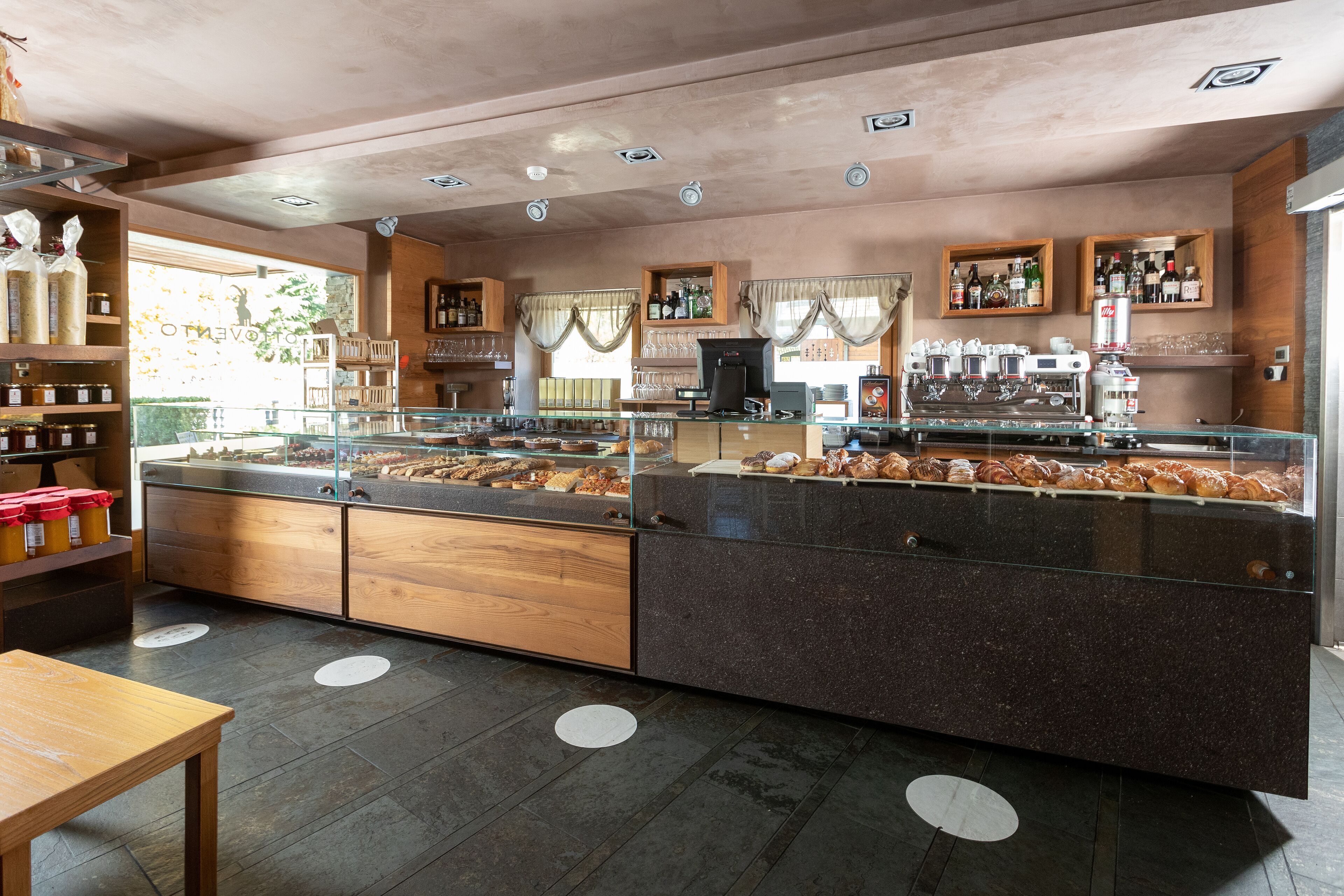Mostrador de panadería con pasteles, una máquina de café y estantes con botellas.