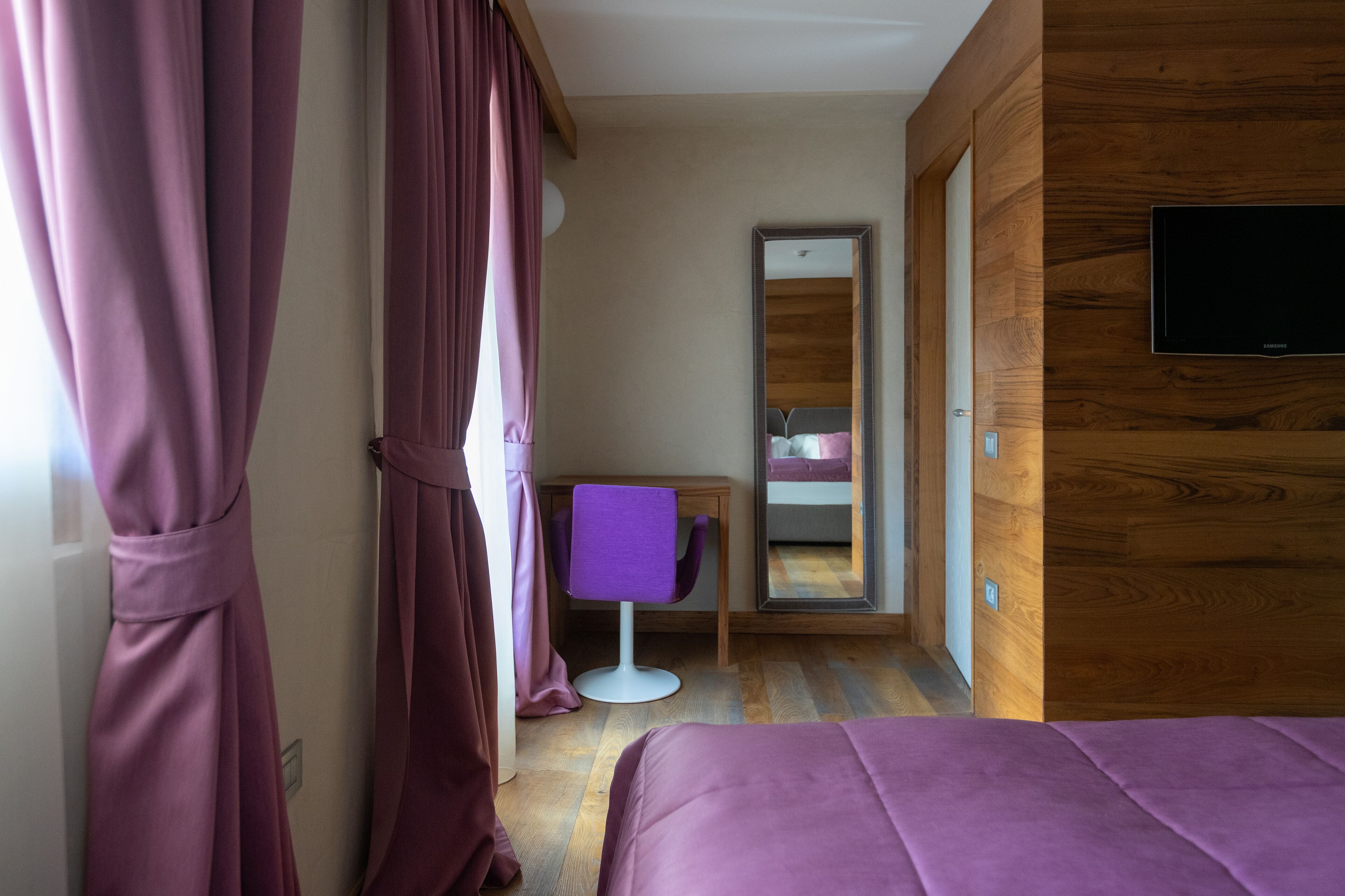 A modern hotel room with a wooden accent wall, a bed, a desk with a chair, and a television.
