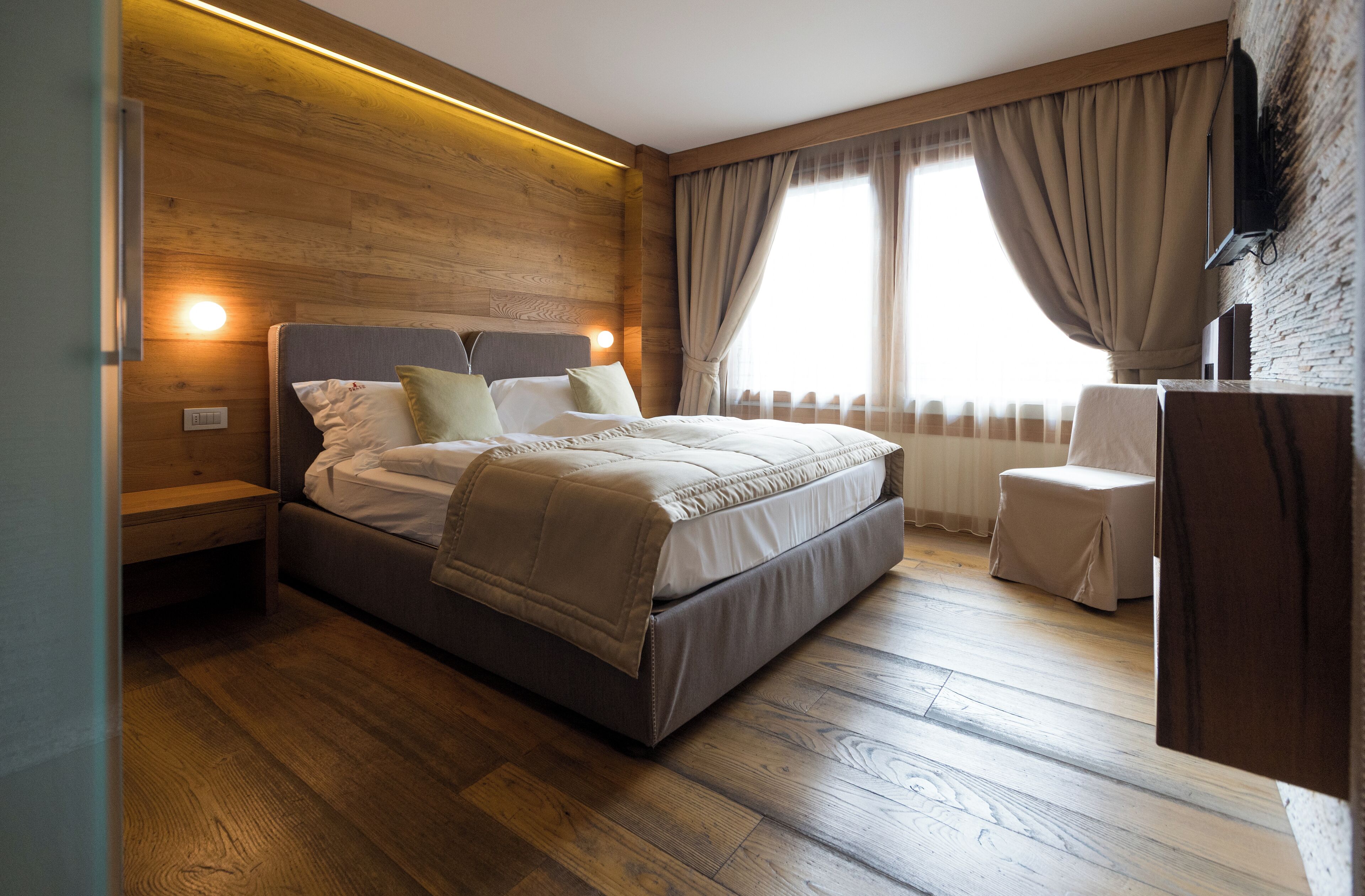 Une chambre à coucher moderne avec une tête de lit en bois, un lit recouvert d’une couette beige, une table de chevet et un petit coin salon avec une chaise et une table.