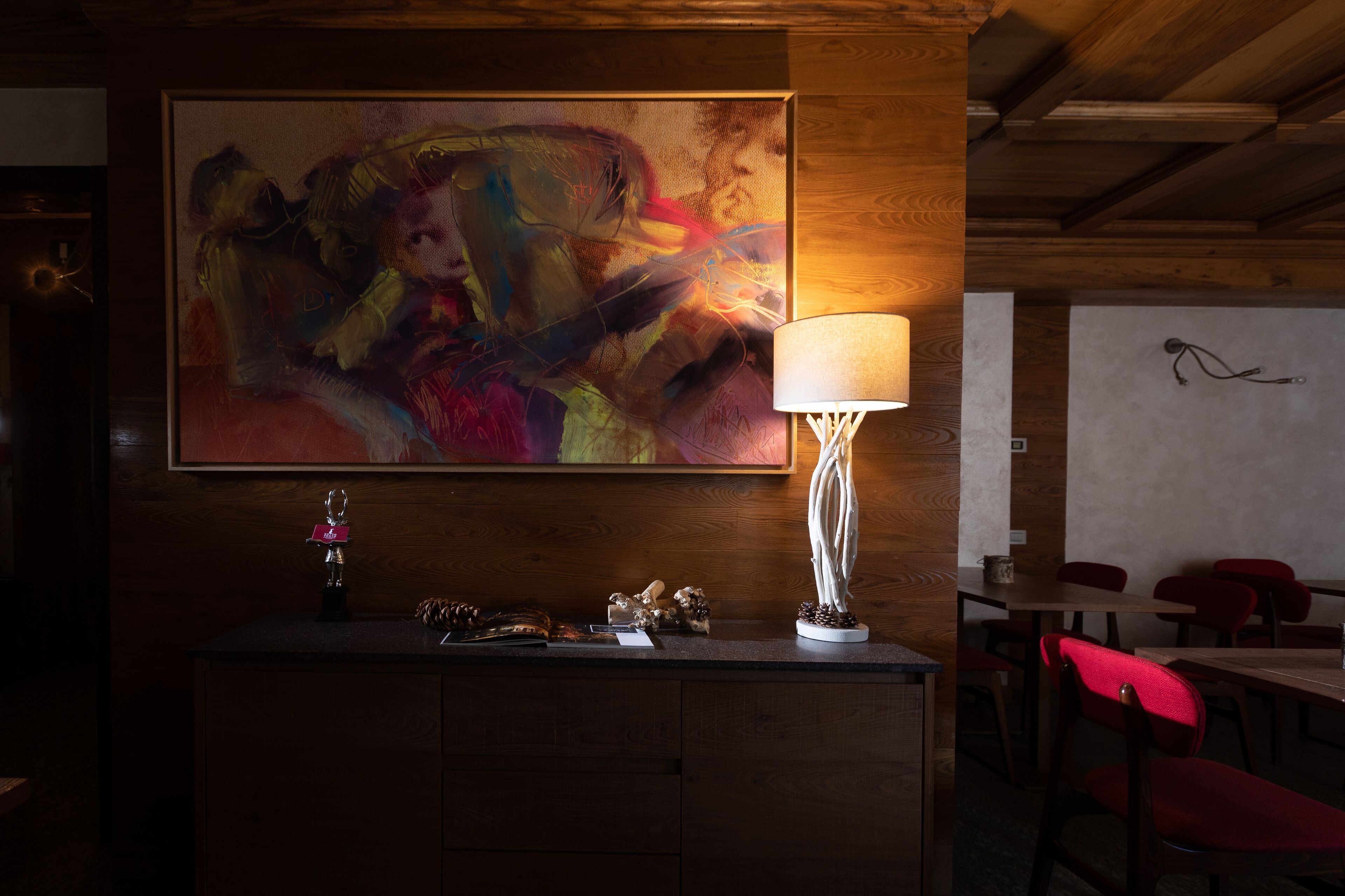 A wooden interior with a large abstract painting, a table lamp, and red chairs.