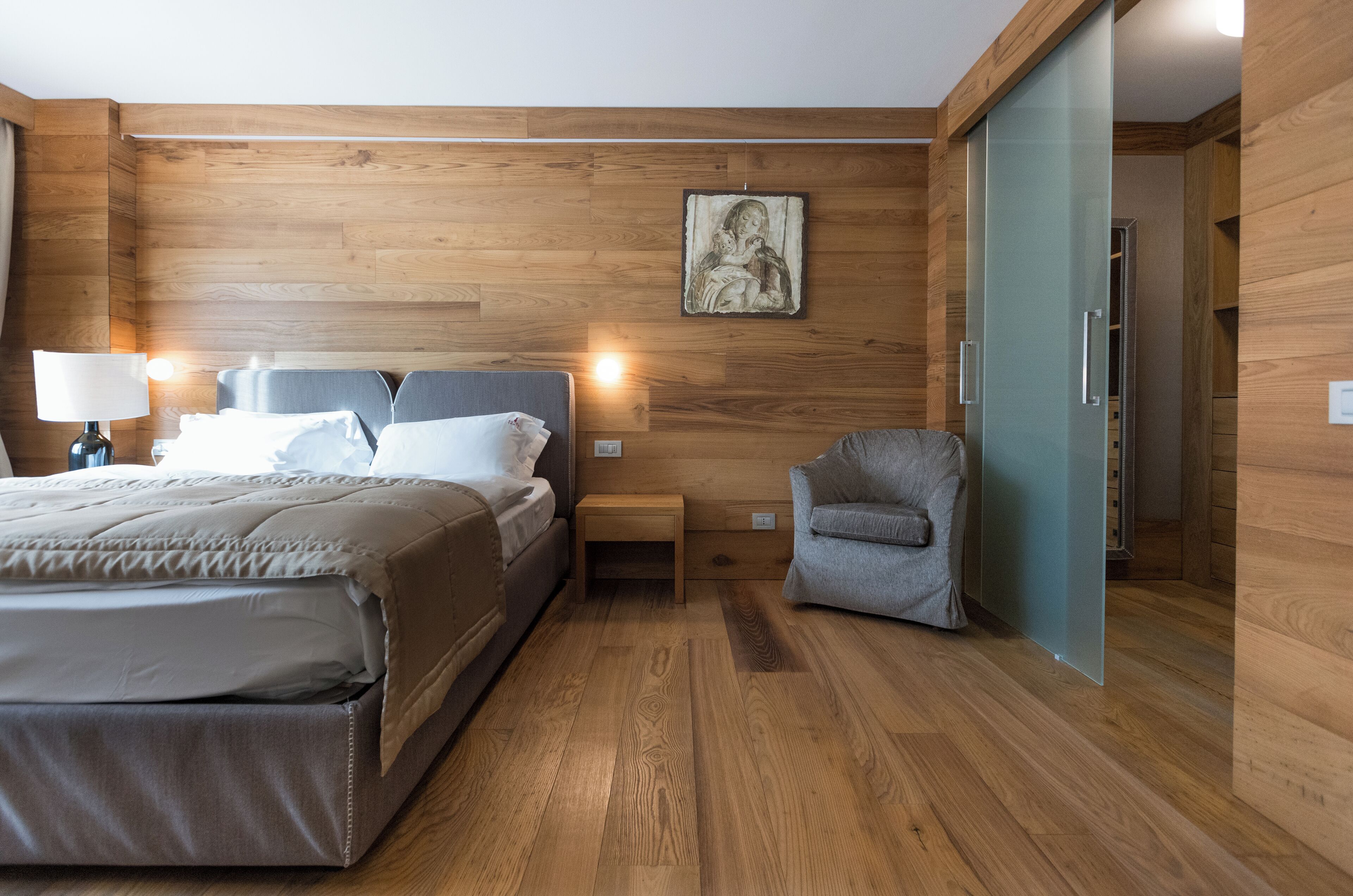 A modern hotel room with wooden walls, a bed with a grey headboard, a grey armchair, a bedside table with a lamp, and a framed picture on the wall.
