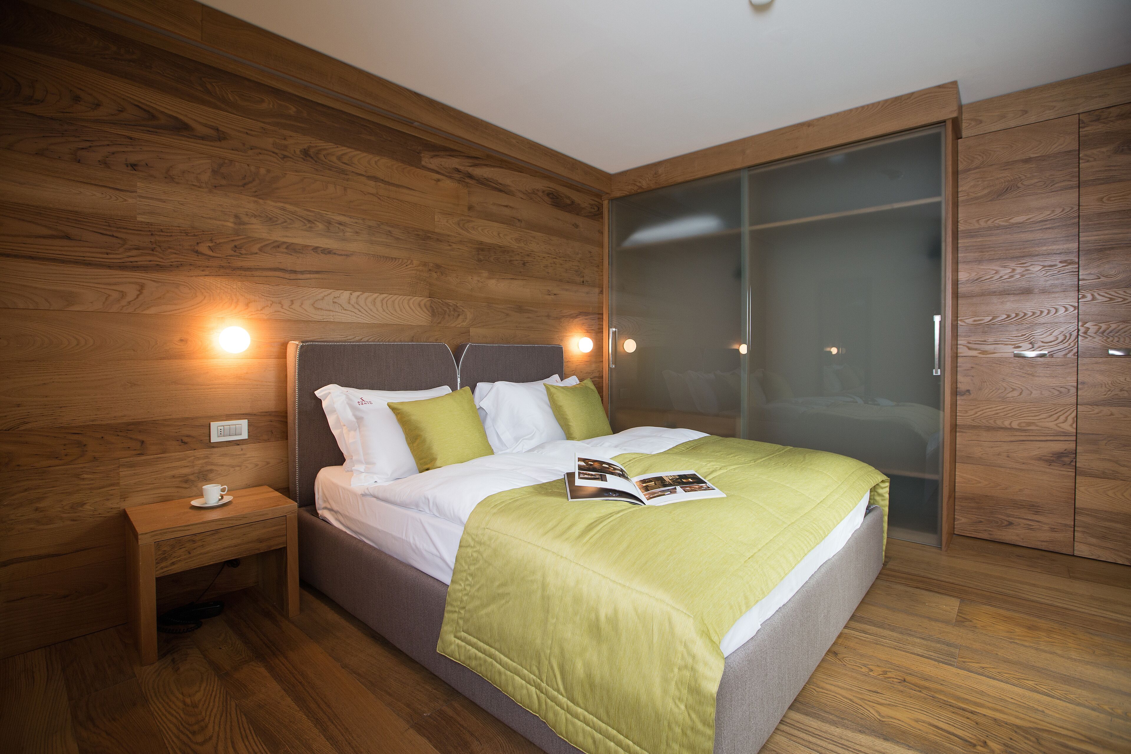 A modern bedroom with a wooden floor, a bed with a green blanket, a bedside table with a cup, and a wardrobe with a glass door.