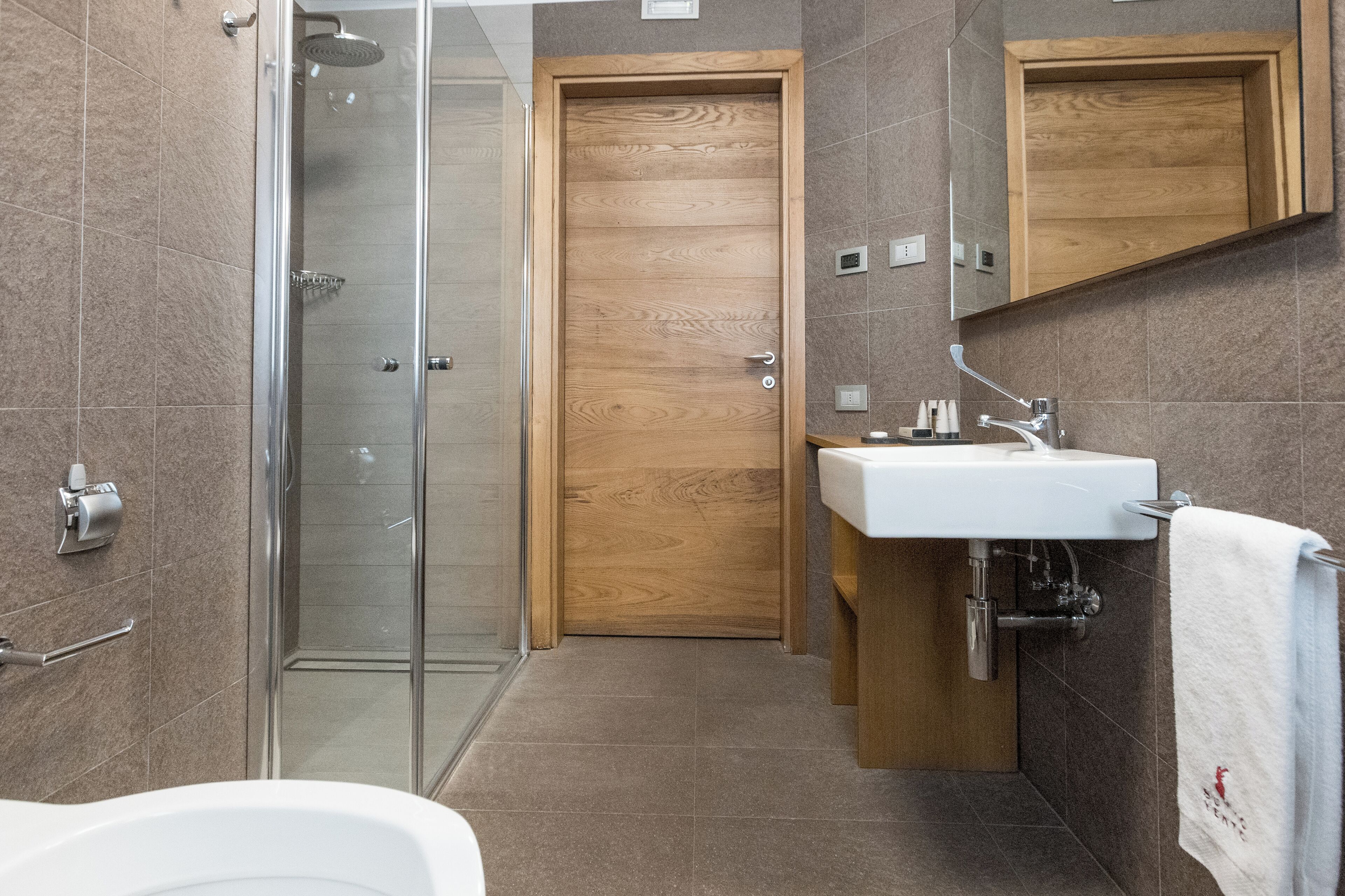 Un baño moderno con cabina de ducha de vidrio, puerta de madera, lavabo blanco y una toalla colgada en un perchero.