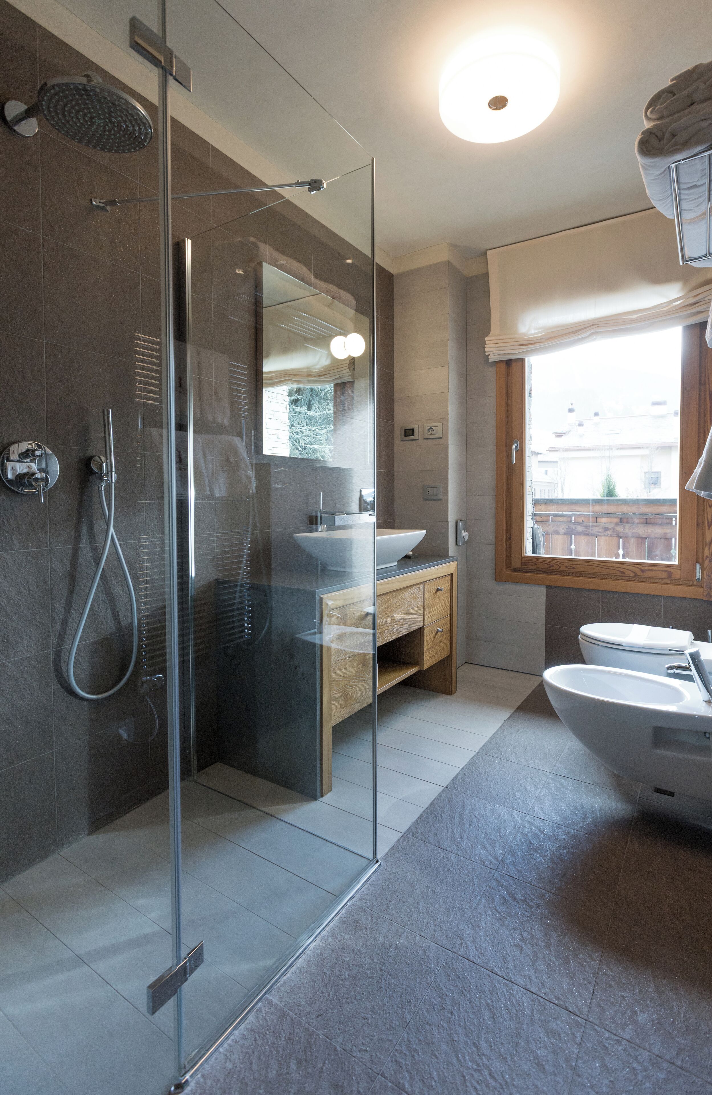 Un bagno moderno con doccia vetrata, mobile lavabo in legno, lavandino bianco e una grande finestra.