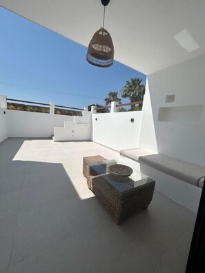 A rooftop terrace with a white wall, a hanging light fixture, a wicker sofa, and a glass table.